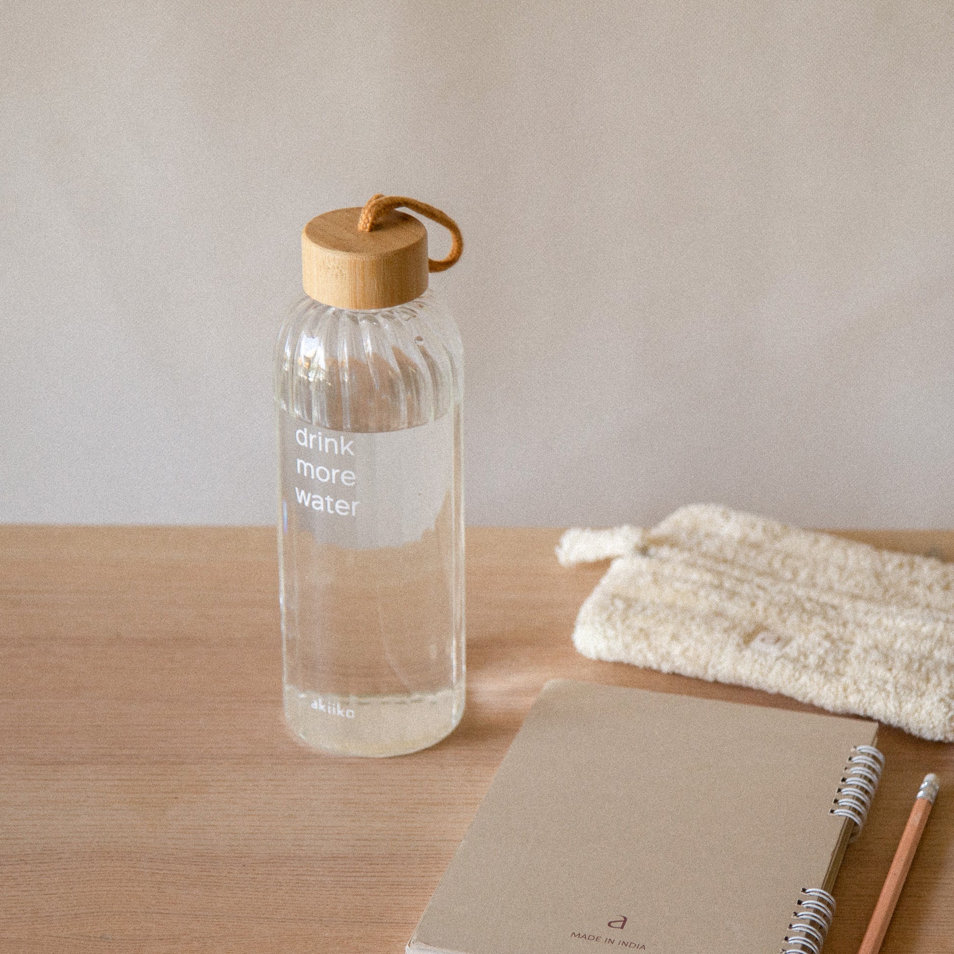 Transparent ribbed glass water bottle with “Drink More Water” text, bamboo lid, and rope handle, 750 ml.