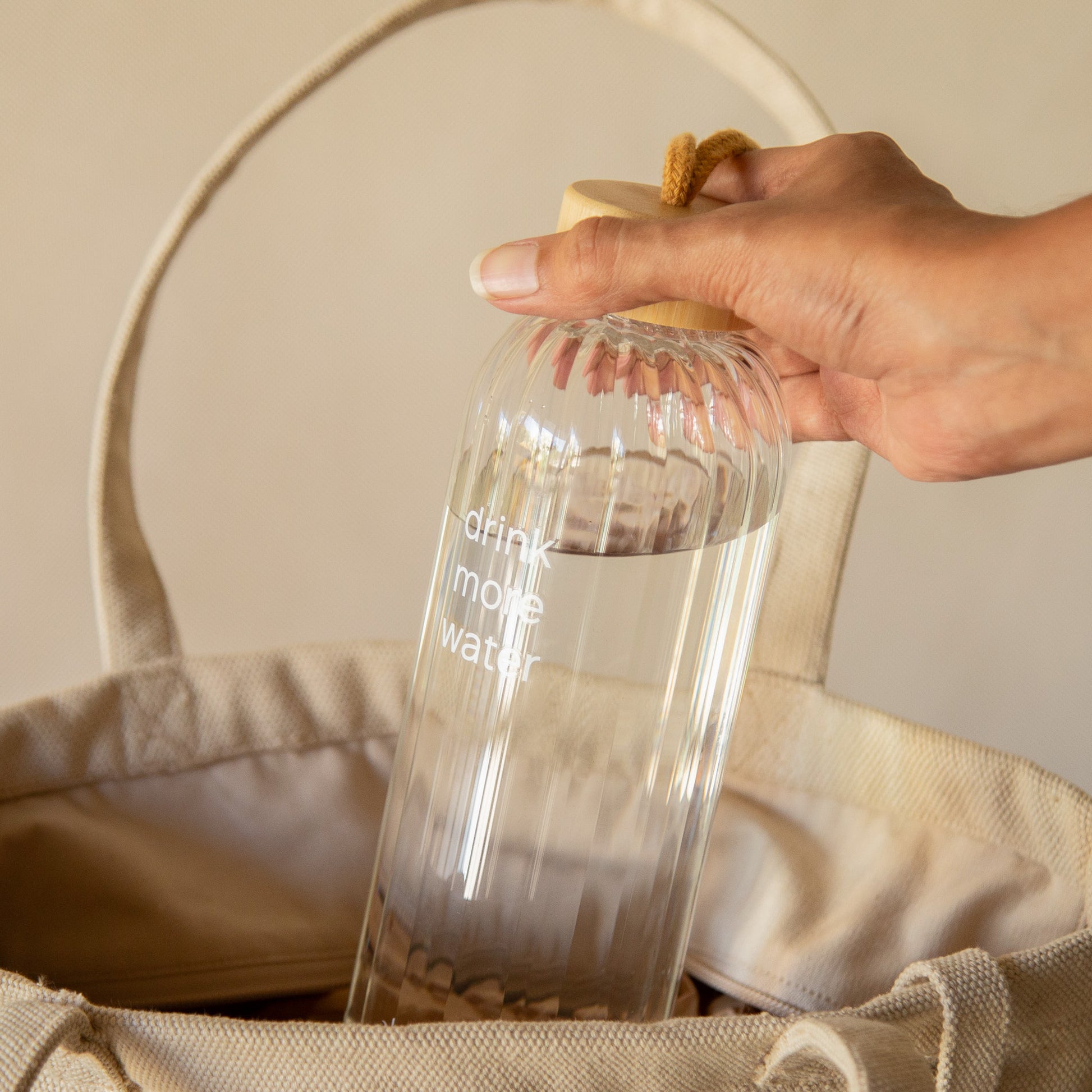 Transparent ribbed glass water bottle with “Drink More Water” text, bamboo lid, and rope handle, 750 ml.