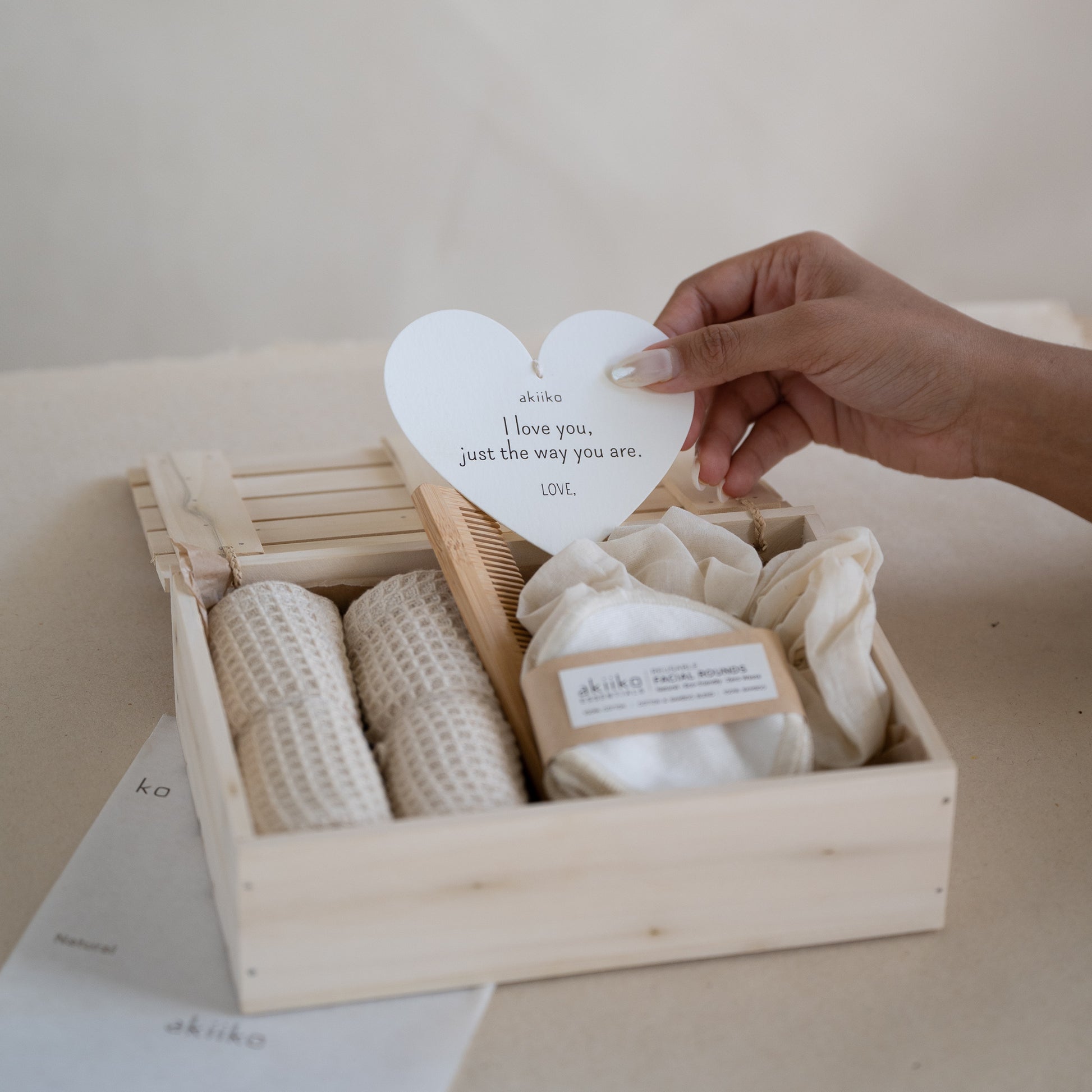 Wooden gift box containing a self-care set with two waffle hand towels, Sutachi scrunchie, six reusable facial rounds, bamboo comb, and a message card.