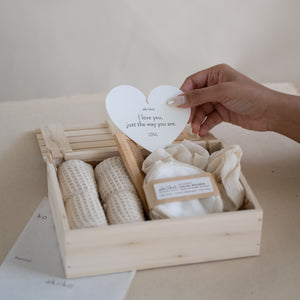 Wooden gift box containing a self-care set with two waffle hand towels, Sutachi scrunchie, six reusable facial rounds, bamboo comb, and a message card.