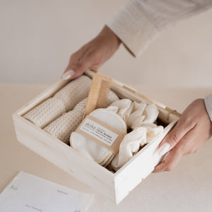 Wooden gift box containing a self-care set with two waffle hand towels, Sutachi scrunchie, six reusable facial rounds, bamboo comb, and a message card.